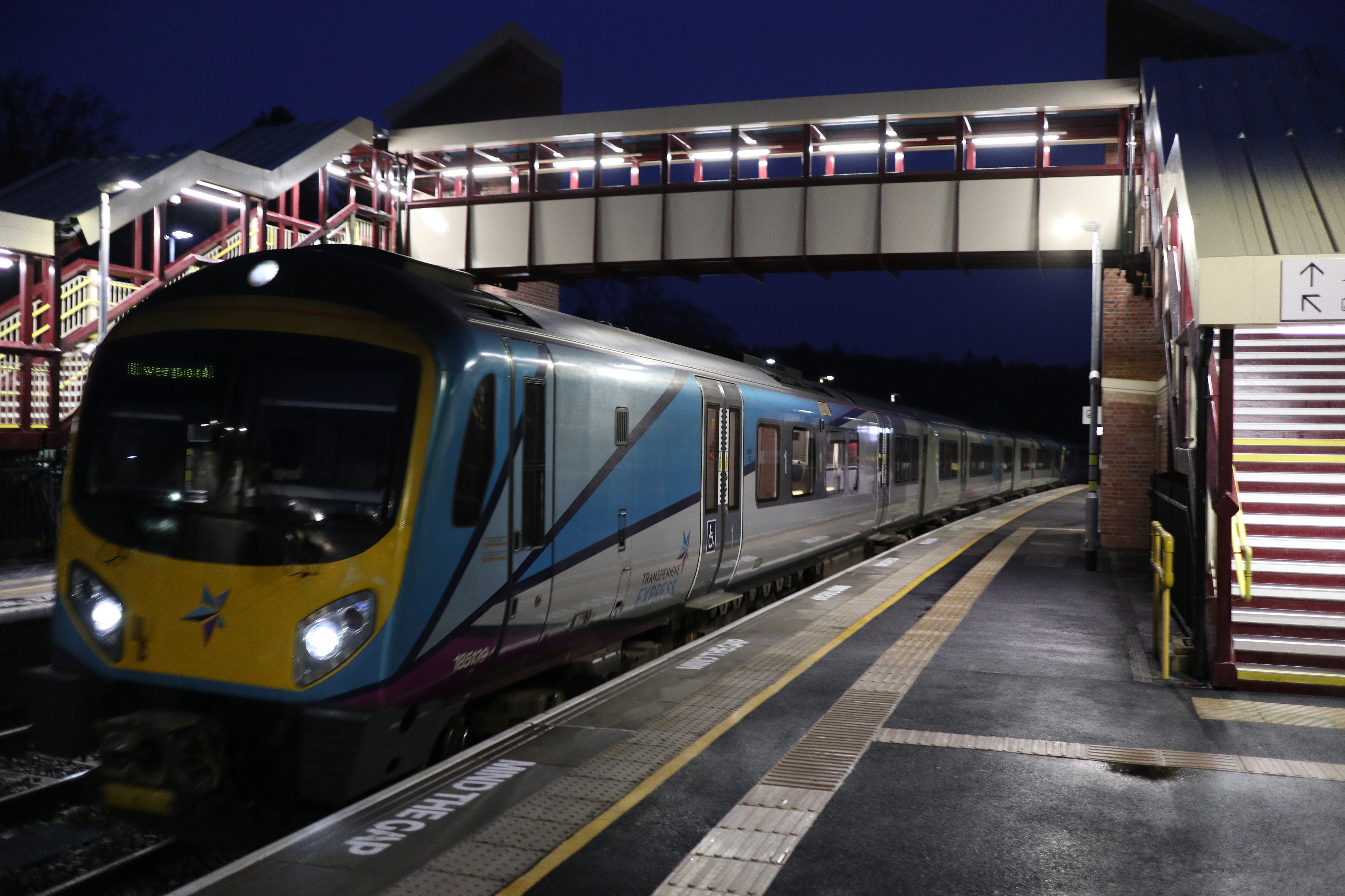 Photo of a passenger train passing under a pedestrian bridge that spans across platforms at a train station