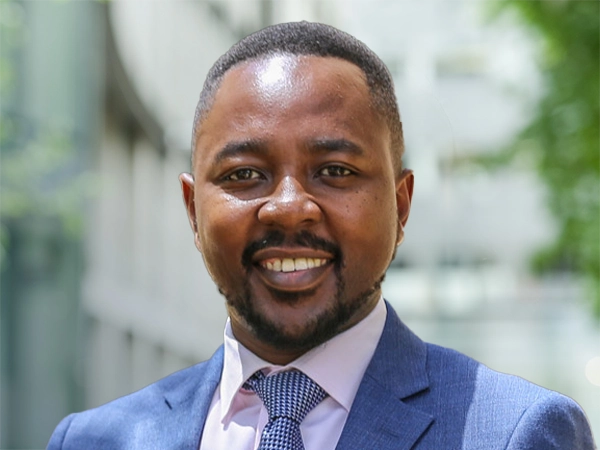 Robert Gichohi, Country Director, standing outside in front of an outdoor urban setting, featuring modern architectural elements with clean lines. There are hints of greenery, suggesting some trees or plants.