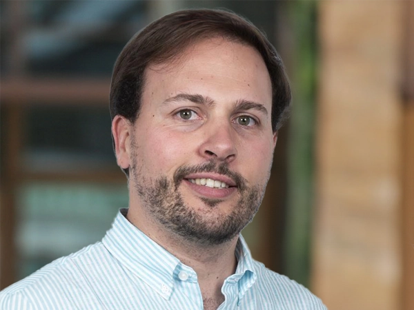 Portrait of Guillermo Martinez, Director, Industrial, Science and Technology, outside in front of a blurred brown background featuring a building and mix of natural and urban elements.