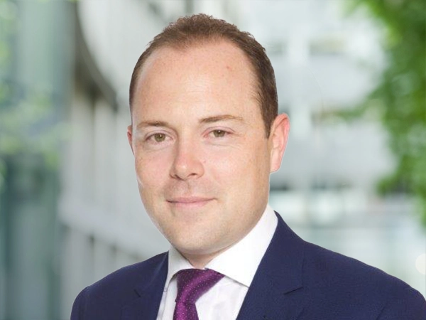 Portrait of Adam Bywater, Director, standing outside in front of an outdoor urban setting, featuring modern architectural elements with clean lines. There are hints of greenery, suggesting some trees or plants.