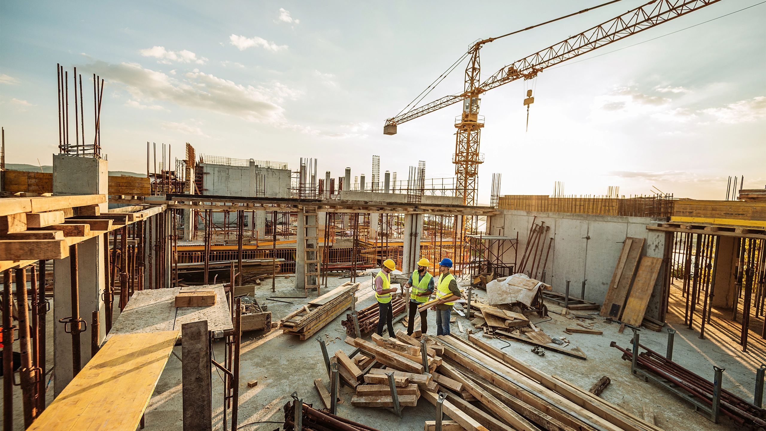 Three male construction workers discussing plans on the floor of a building under construction with a crane in background skyline.