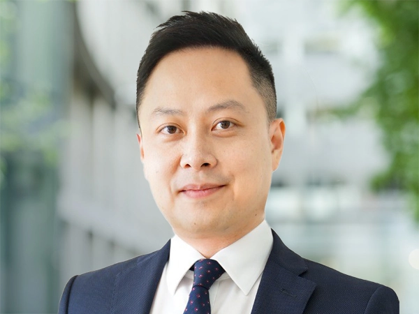 Daniel Cheung, Strategic Lead, Hong Kong & Macau, and Head of Infrastructure, Northeast Asia, standing outside in front of an outdoor urban setting, featuring modern architectural elements with clean lines. There are hints of greenery, suggesting some trees or plants.