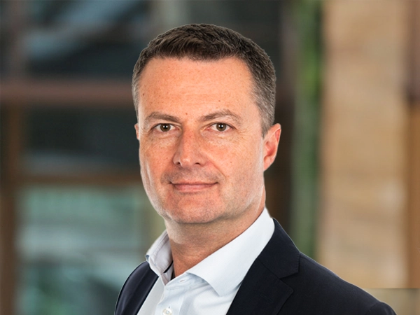Portrait of Mark Rogers, Director, UK, outside in front of a blurred brown background featuring a building and mix of natural and urban elements.