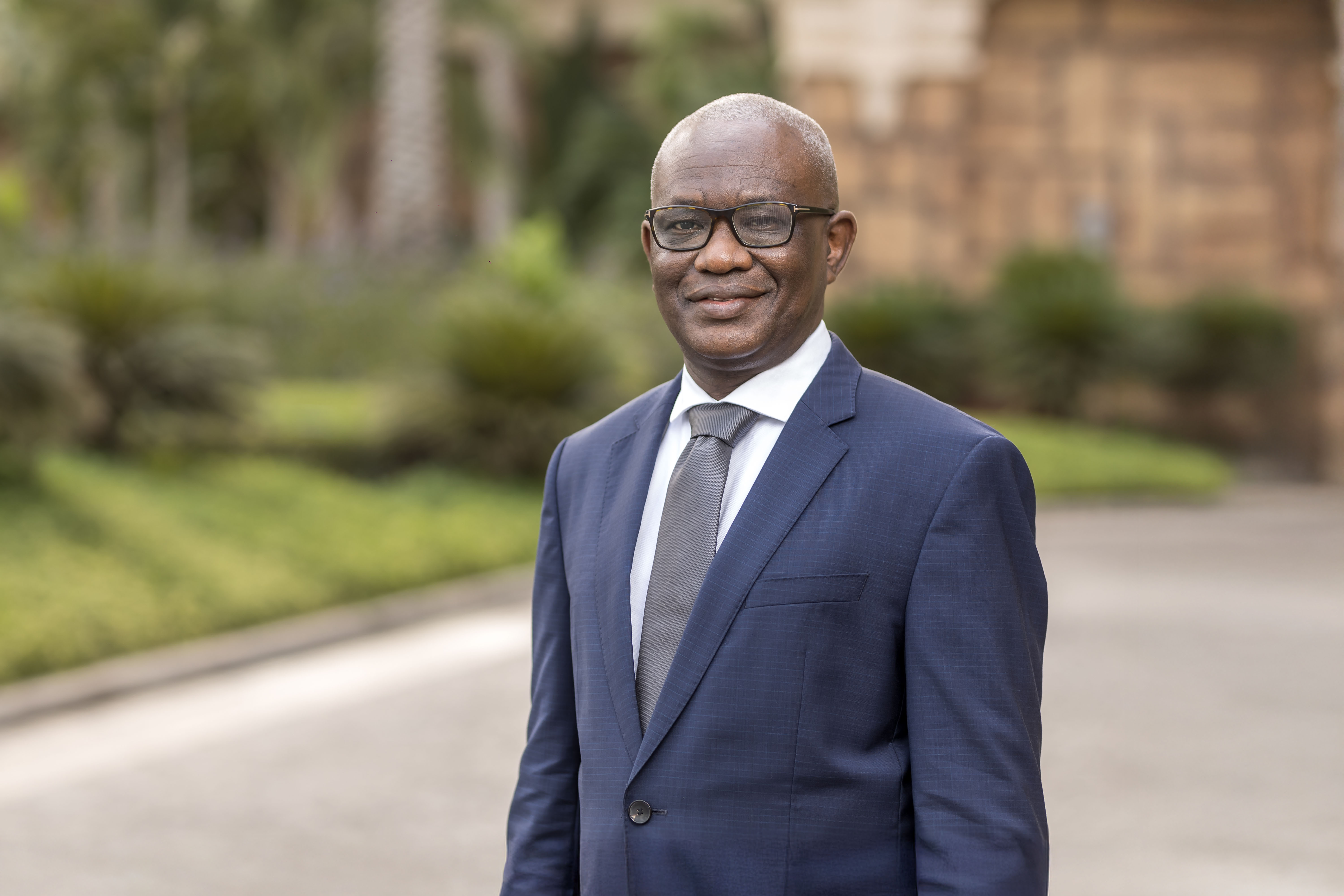 Portrait of Segun Faniran, Forensic Planning and Delay, Middle East, outside in front of grass and bushes with tall buildings behind.