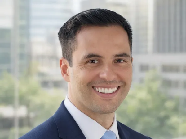 Portrait of Andrew Pollard, Director, standing against office window with tall city views behind and green trees in background.