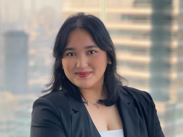 Portrait of Nanda Angelina, Marketing and Communications Coordinator, Asia, Smiling in office space against window, where blurry figures of other offices buildings are shown.