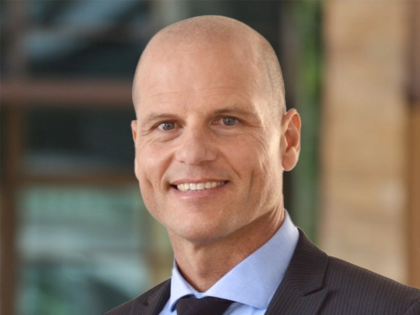 Brad Moolman, Associate Director, outside in front of a blurred brown background featuring a building and mix of natural and urban elements.