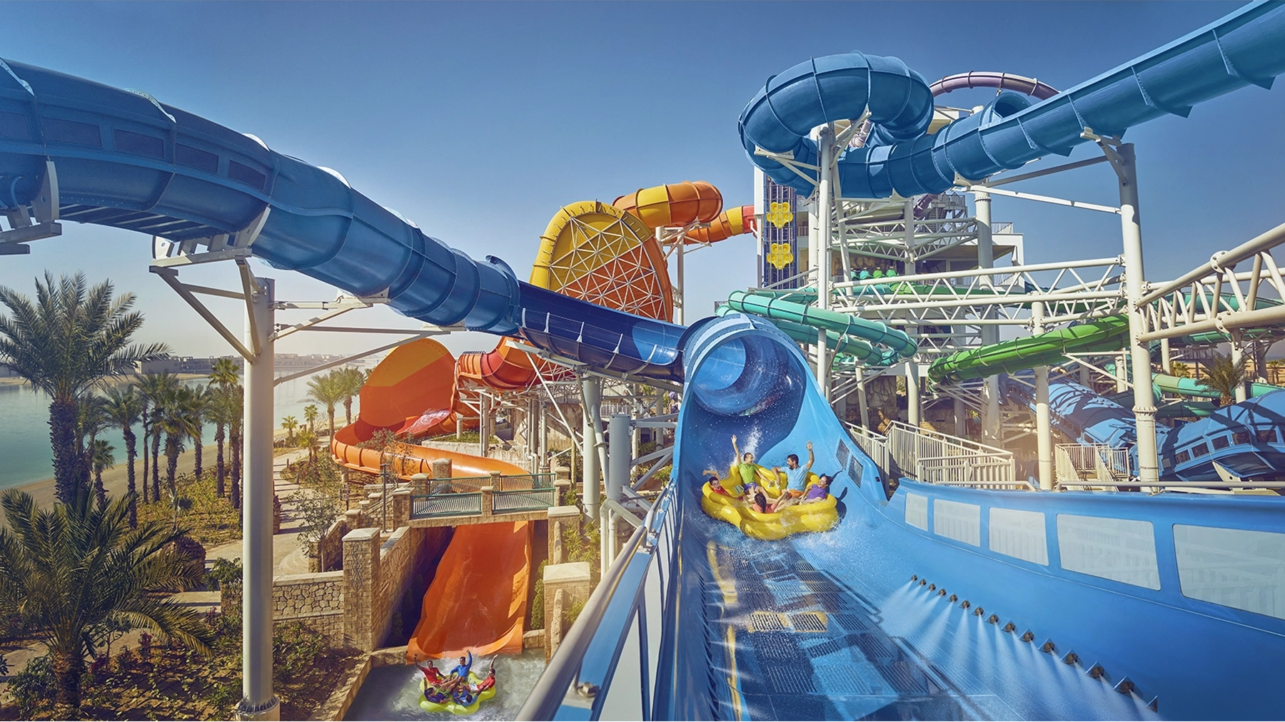 Big water park with lots of slides, featuring family on water float going down big blue water slide with heir arms raised in the air