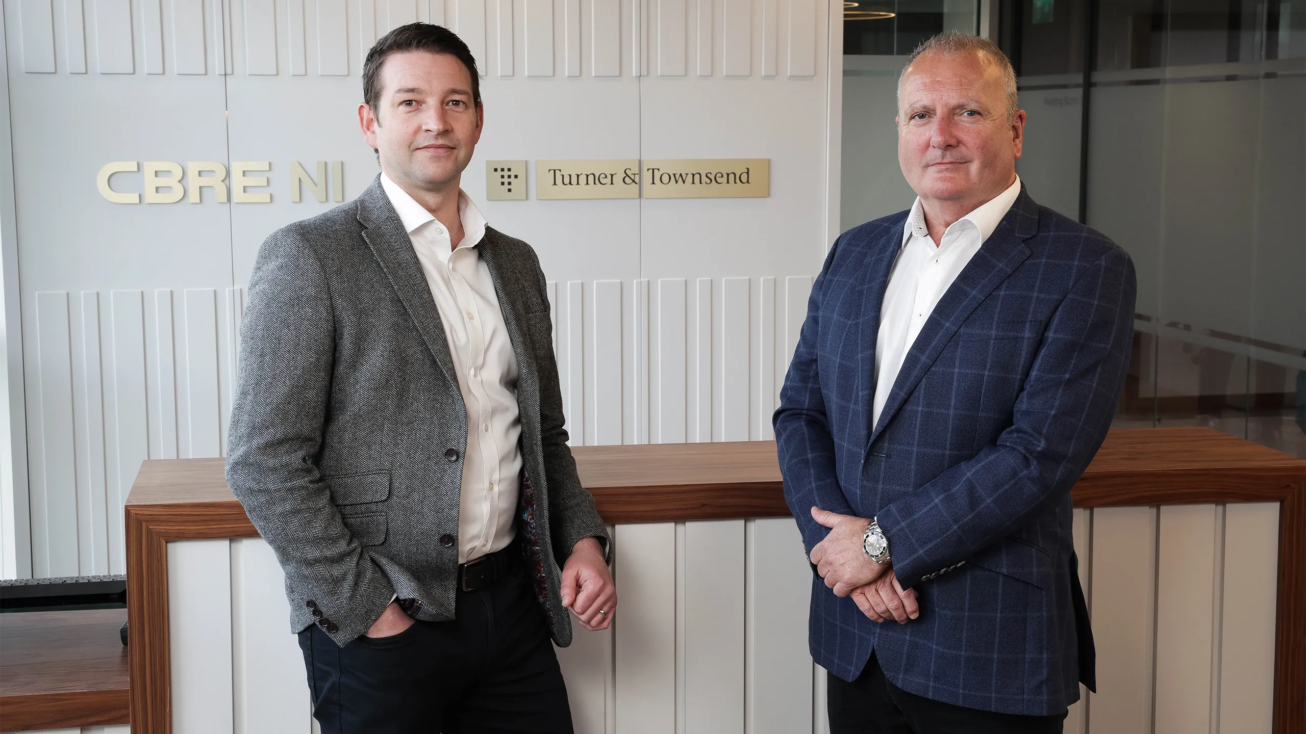 Two business men in suits, standing in front of grey back ground with gold CBRE and Turner & Townsend logo.