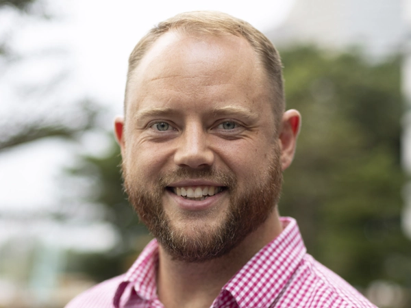 Liam Bray, Director, Portfolio Management, standing outside. There are hints of greenery, suggesting some trees or plants.