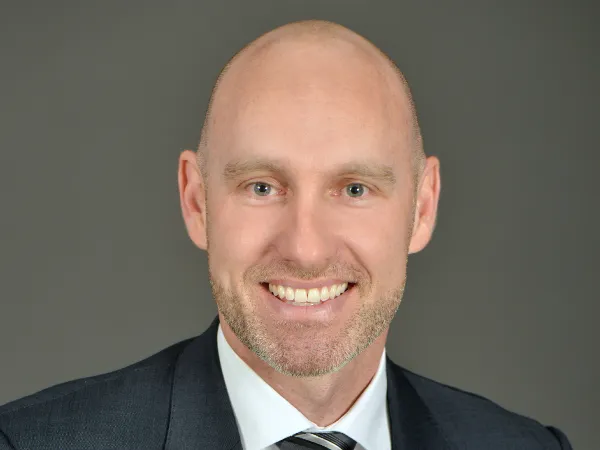 Portrait of Mark Utting, Managing Director, Switzerland, smiling in a suit against dark grey background.
