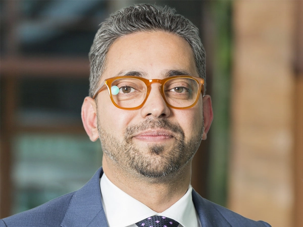 Satch Bhattacharjee, Key Client Lead, Australia, outside in front of a blurred brown background featuring a building and mix of natural and urban elements.