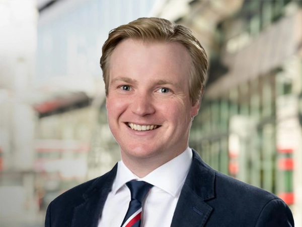 Portrait of George Highfield, Real Estate Advisory, Data & Digital – Principal Consultant, standing outside in front of buildings featuring an urban cityscape with a mix of architectural styles. The scene includes some windows and red features, adding a touch of color.