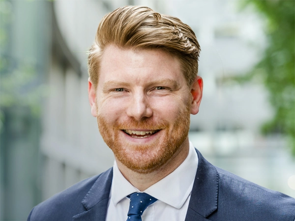 Guy Beaumont, Digital Lead, Infrastructure, standing outside in front of an outdoor urban setting, featuring modern architectural elements with clean lines. There are hints of greenery, suggesting some trees or plants.