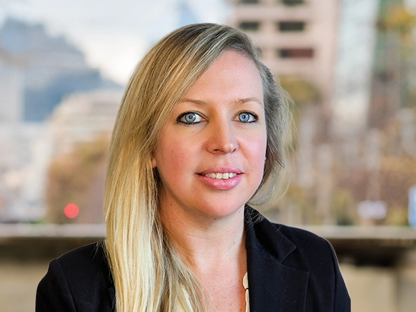 portrait of Caitlin Adair, Director, with long blonde hair, standing outside featuring an urban cityscape with tall buildings and a mix of architectural styles. The scene includes some trees with autumn foliage, adding a touch of color. 