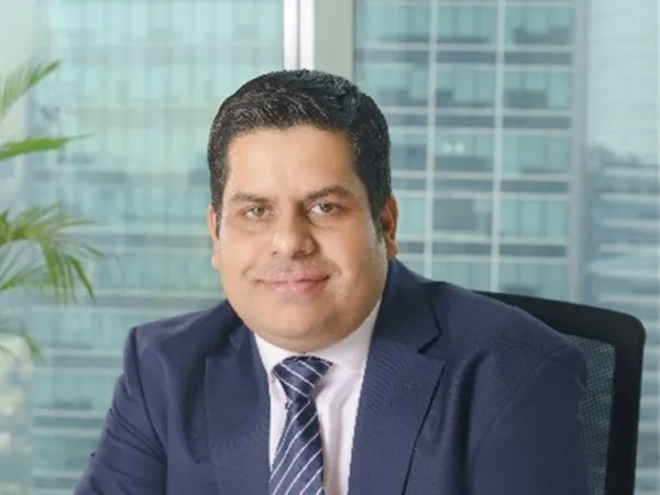 Sumit Mukherjee, Director, standing outside in front of buildings featuring an urban cityscape with a mix of architectural styles. The scene includes some windows and red features, adding a touch of color.