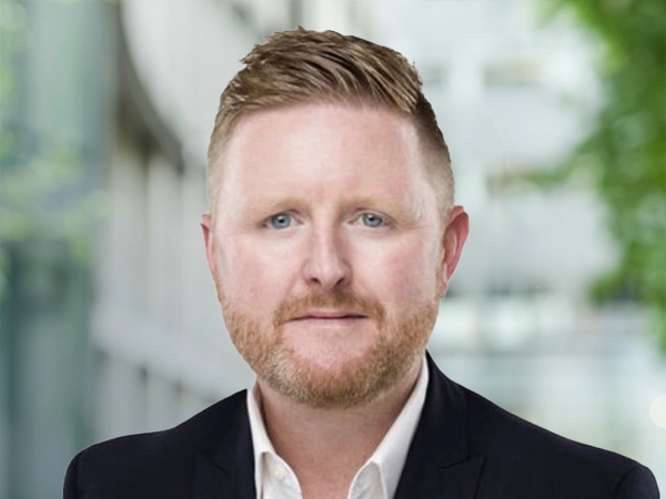 Sean Hyland, Project Director, standing outside in front of an outdoor urban setting, featuring modern architectural elements with clean lines. There are hints of greenery, suggesting some trees or plants.