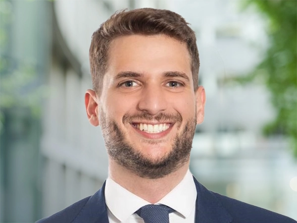Rodrigo Martini, Senior Project Manager, standing outside in front of an outdoor urban setting, featuring modern architectural elements with clean lines. There are hints of greenery, suggesting some trees or plants.