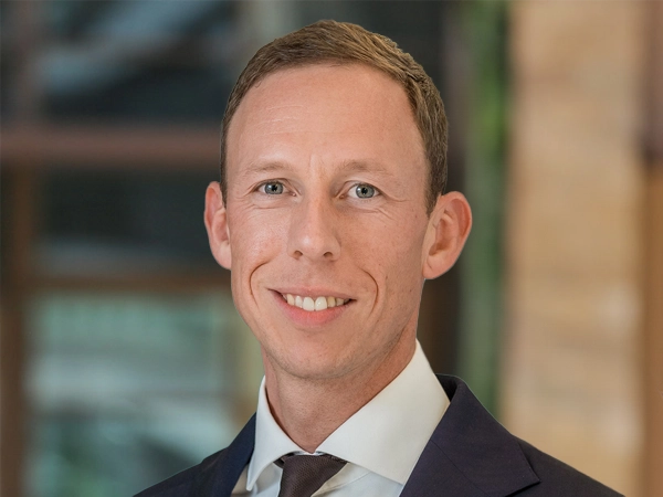 Portrait of Dennis de Wolf, Director, Real Estate, outside in front of a blurred brown background featuring a building and mix of natural and urban elements.