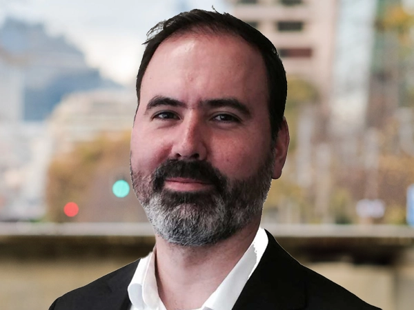 Gonzalo Rosa, Director, standing outside featuring an urban cityscape with tall buildings and a mix of architectural styles. The scene includes some trees with autumn foliage, adding a touch of color. 