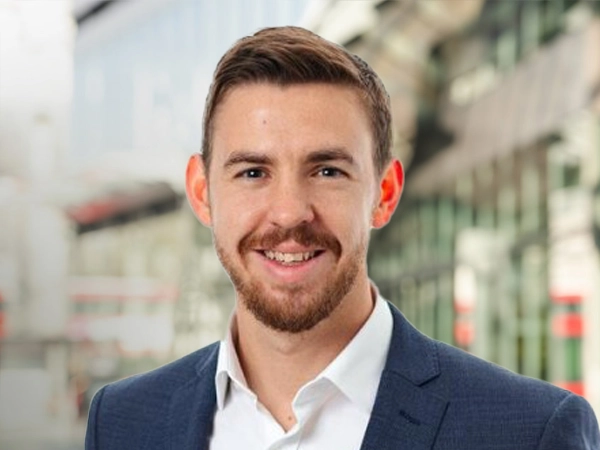 Portrait of Richard Farrelly, Project Director, UK, standing outside in front of buildings featuring an urban cityscape with a mix of architectural styles. The scene includes some windows and red features, adding a touch of color.