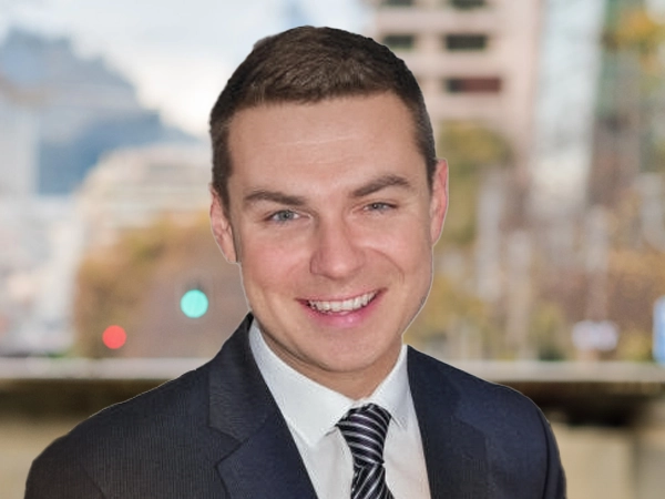 Portrait of Colin McBride, Director of Real Estate, Hong Kong and Macau, Standing outside featuring an urban cityscape with tall buildings and a mix of architectural styles. The scene includes some trees with autumn foliage, adding a touch of color. 