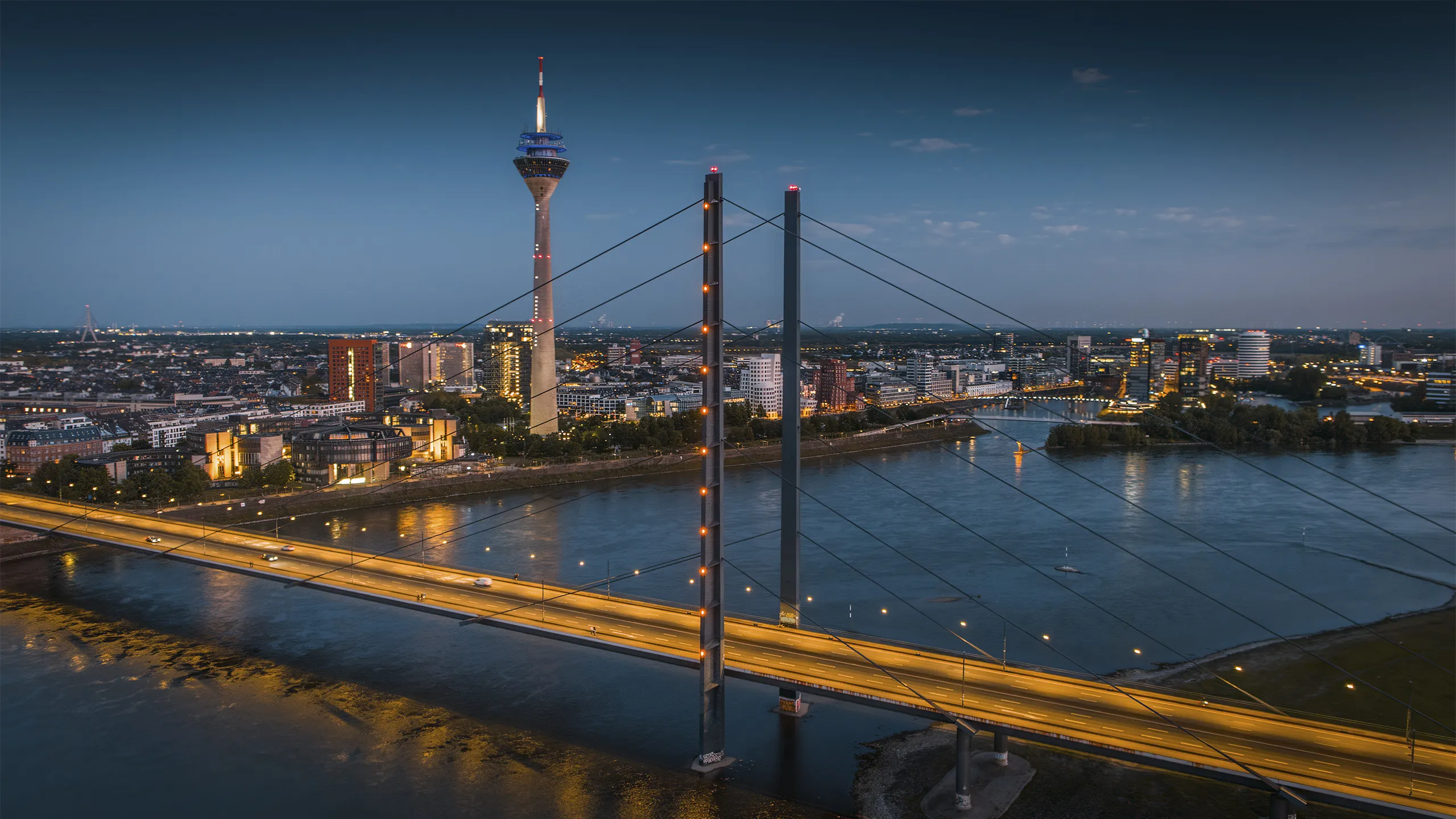Aerial blue-hour view of the Düsseldorf skyline along the Rhine River, featuring the illuminated Rheinkniebrücke leading toward the city center and the Rheinturm rising above modern buildings. City lights reflect on the calm water, creating a clean, contemporary European cityscape. North Rhine–Westphalia, Germany, Europe.