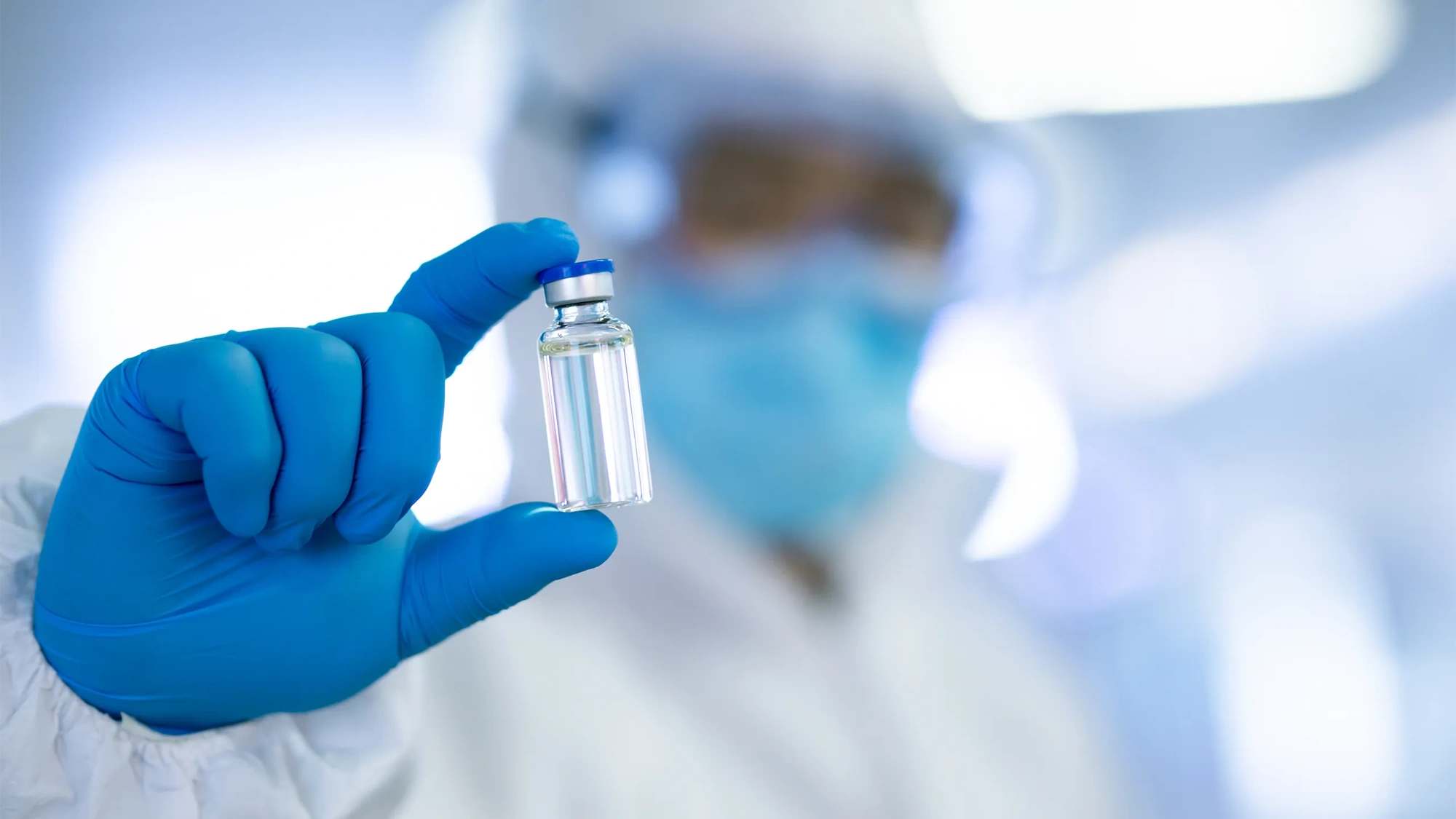 A person dressed in protective laboratory clothing, including blue latex gloves and holding vaccination vile in between their fingers. 