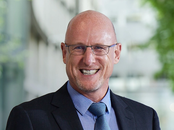 Portrait of John Fowlie, Regional Director, Energy and Natural Resources, standing outside in front of an outdoor urban setting, featuring modern architectural elements with clean lines. There are hints of greenery, suggesting some trees or plants.