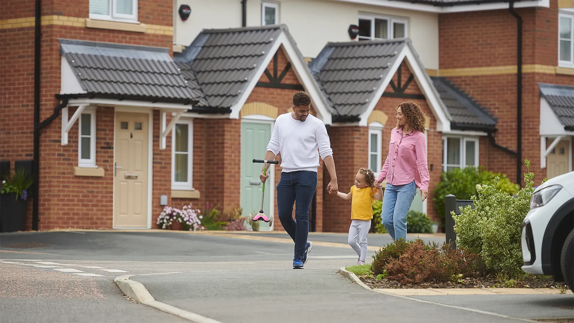 Two adults holding the hand of a child in yellow top outside of housing estate 