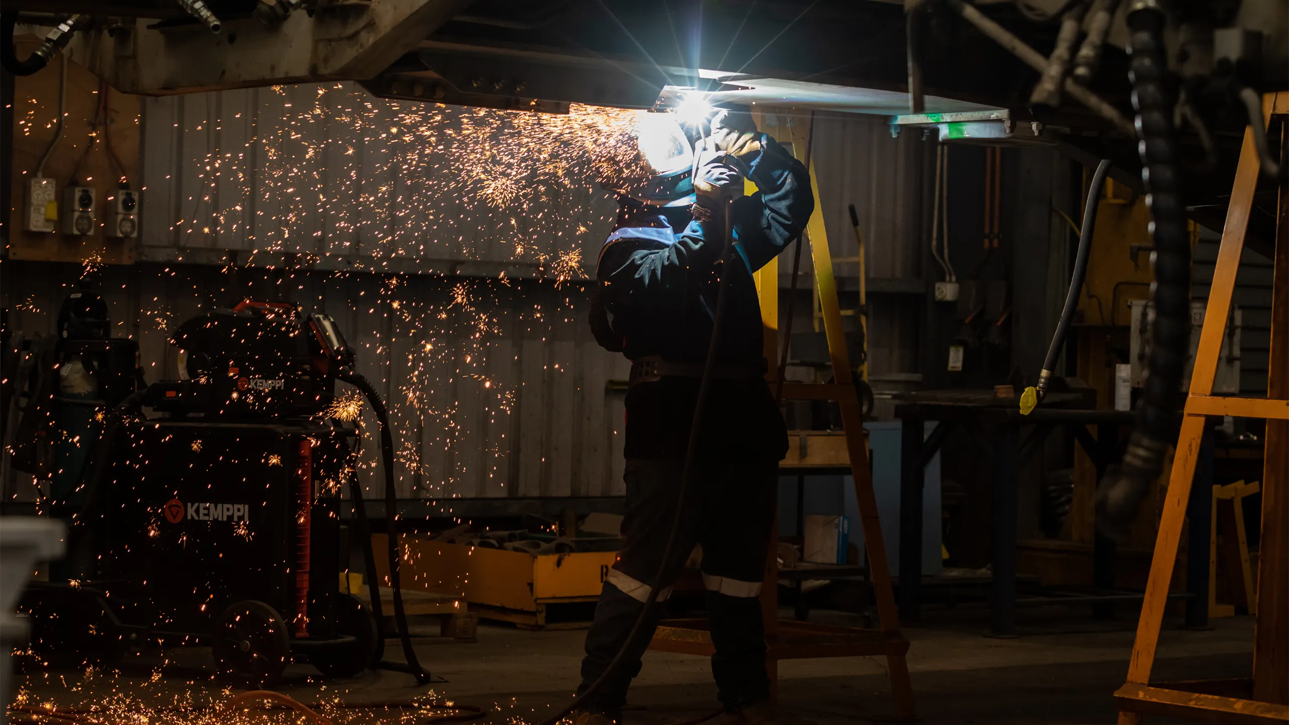 A welder in protective gear, including a welding helmet and gloves, is working in a dimly lit workshop. Bright orange sparks fly outward as they weld metal overhead, illuminating the area around them. Industrial equipment and tools are visible in the background, creating a rugged and workshop-like atmosphere