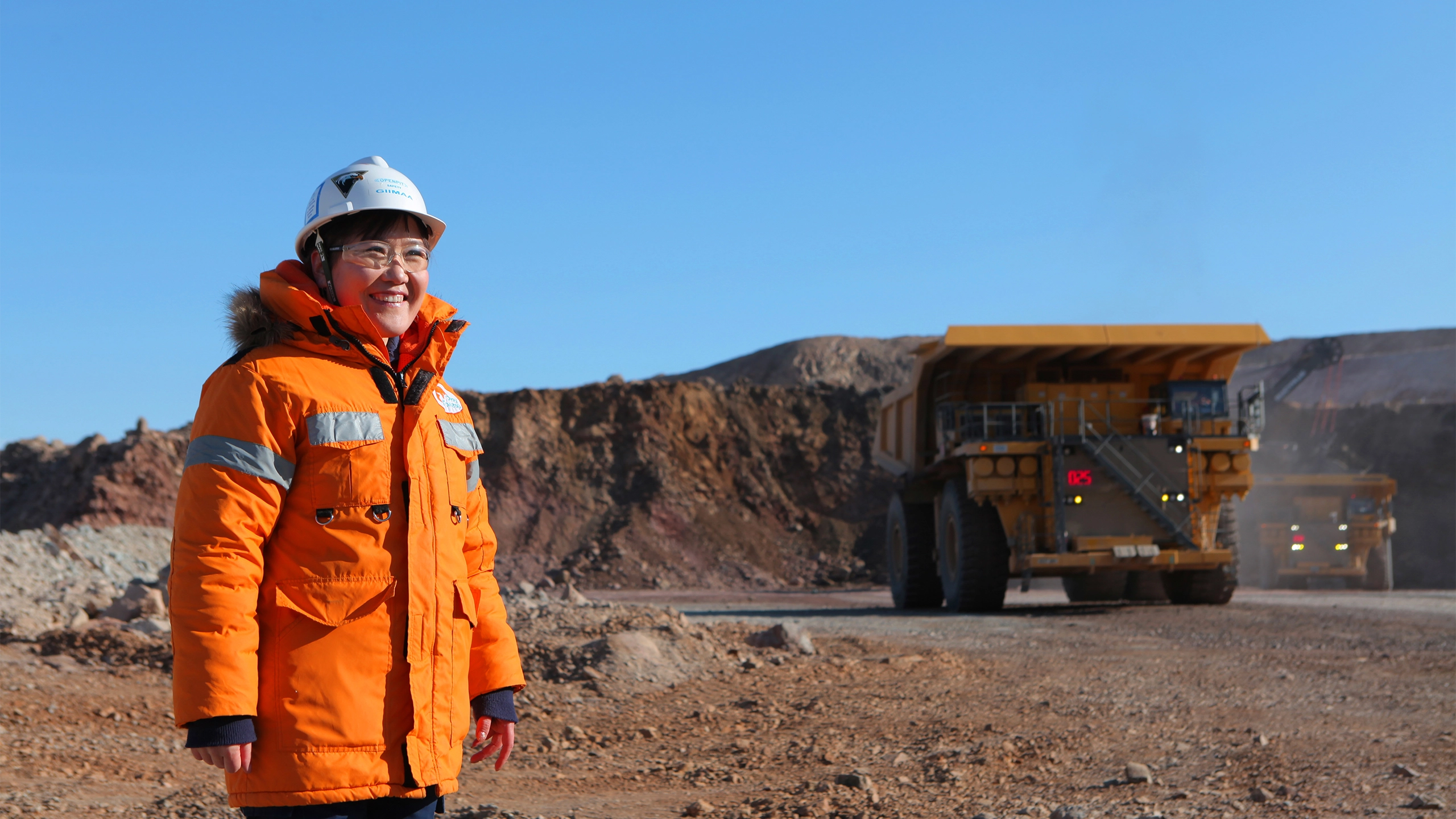 Woman standing in sandy construction site wearing orange hi vis and helmet