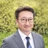 Portrait of Toshio Kosuge, Director, Clean Energy and Transmission, Japan, standing outside against green bushes, in grey suit smiling.