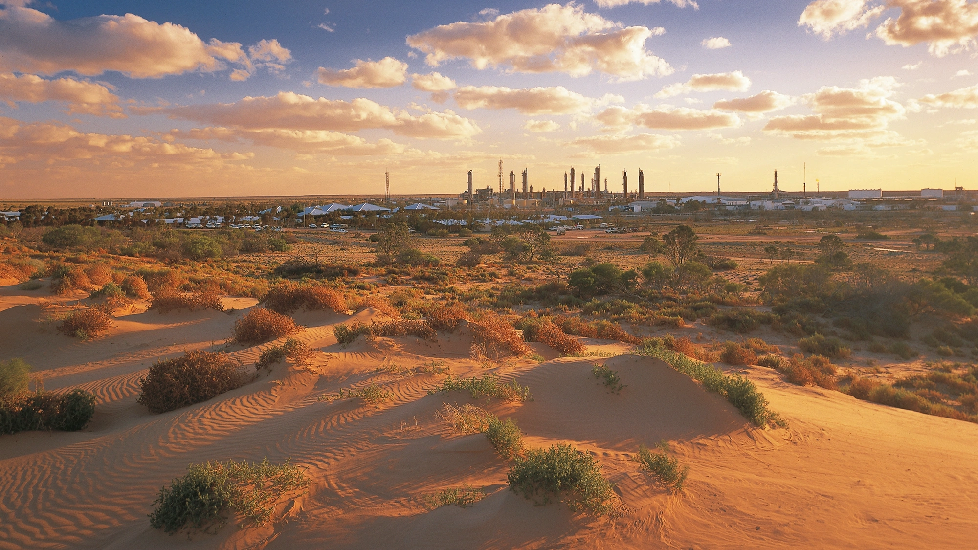 A sandy desert with some tufts of plants in the foreground of the image, with a clean energy plant far away in the distance. 