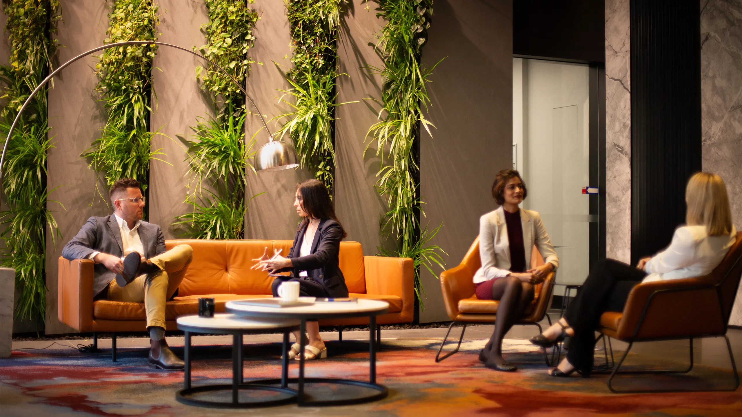 Group of four colleagues sitting separately in pairs on orange sofas in a plant-filled office.
