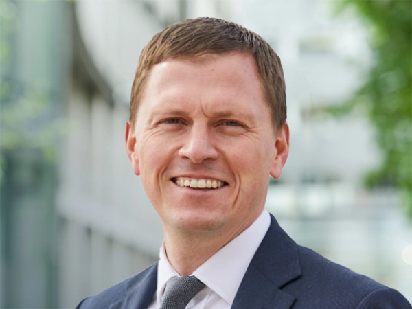 Portrait of Dan Ayley, Global Head of Industrial, Science and Technology, standing outside in front of an outdoor urban setting, featuring modern architectural elements with clean lines. There are hints of greenery, suggesting some trees or plants.