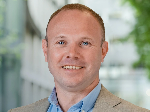 Tim James, Associate Director, standing outside in front of an outdoor urban setting, featuring modern architectural elements with clean lines. There are hints of greenery, suggesting some trees or plants.