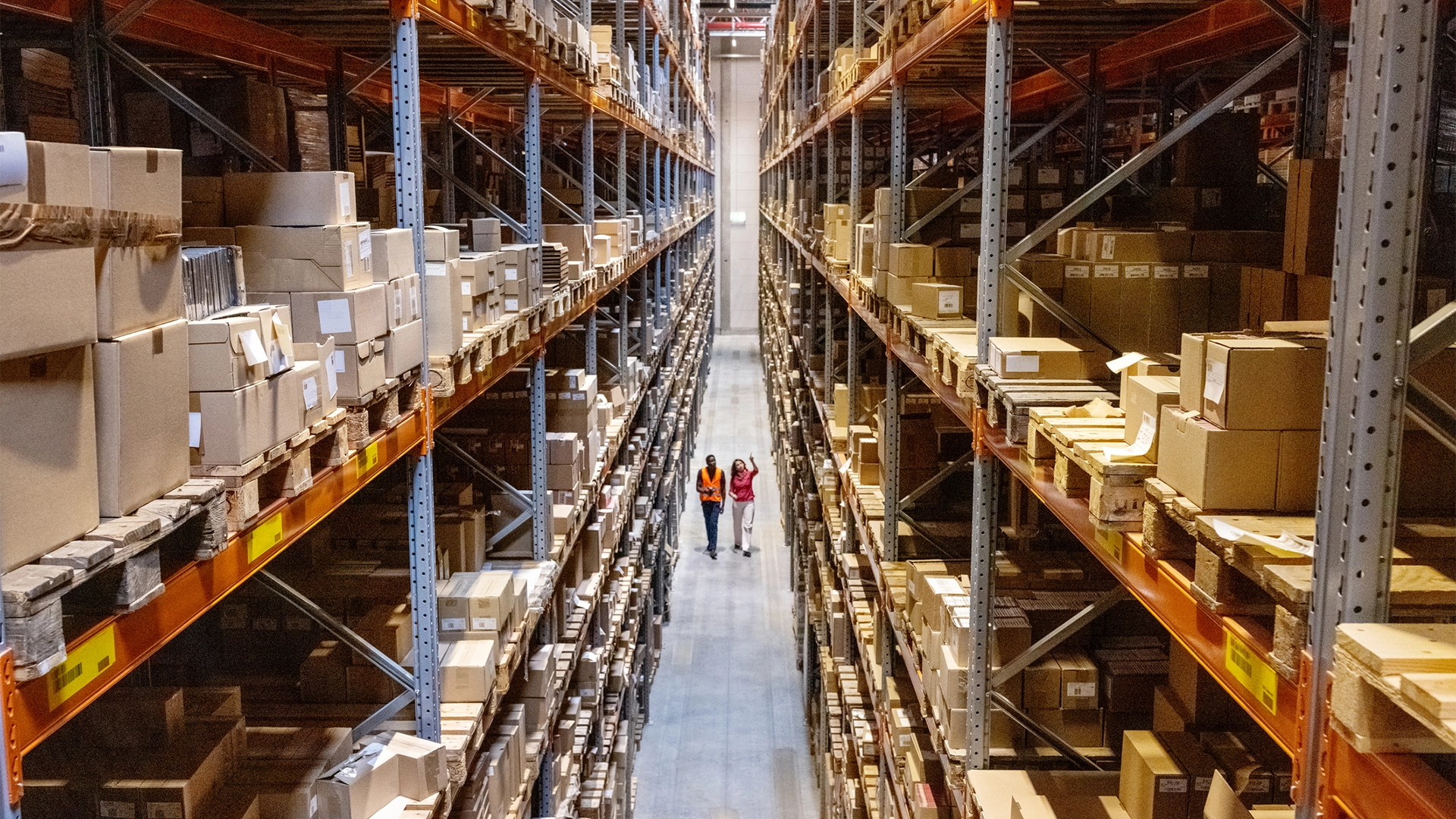 Two employees in high vis jackets walking through warehouse with tall shelves full of products