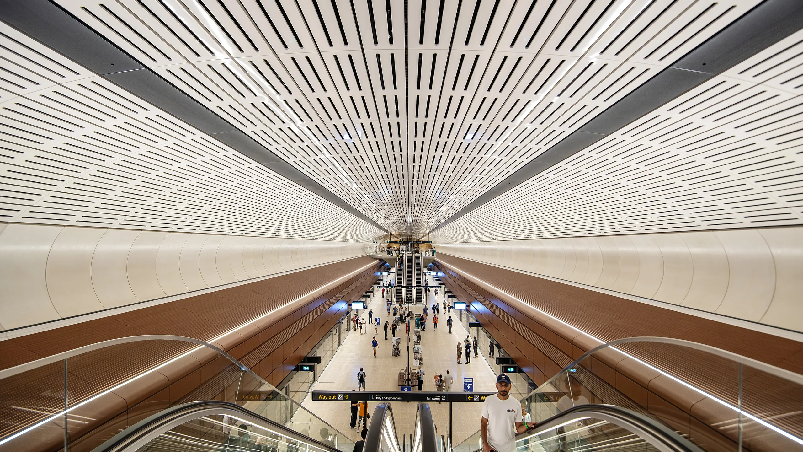 Interior view of Sydney metro modern underground station with a wide ceiling featuring linear ventilation panels, symmetrical architecture and passengers moving through space below.