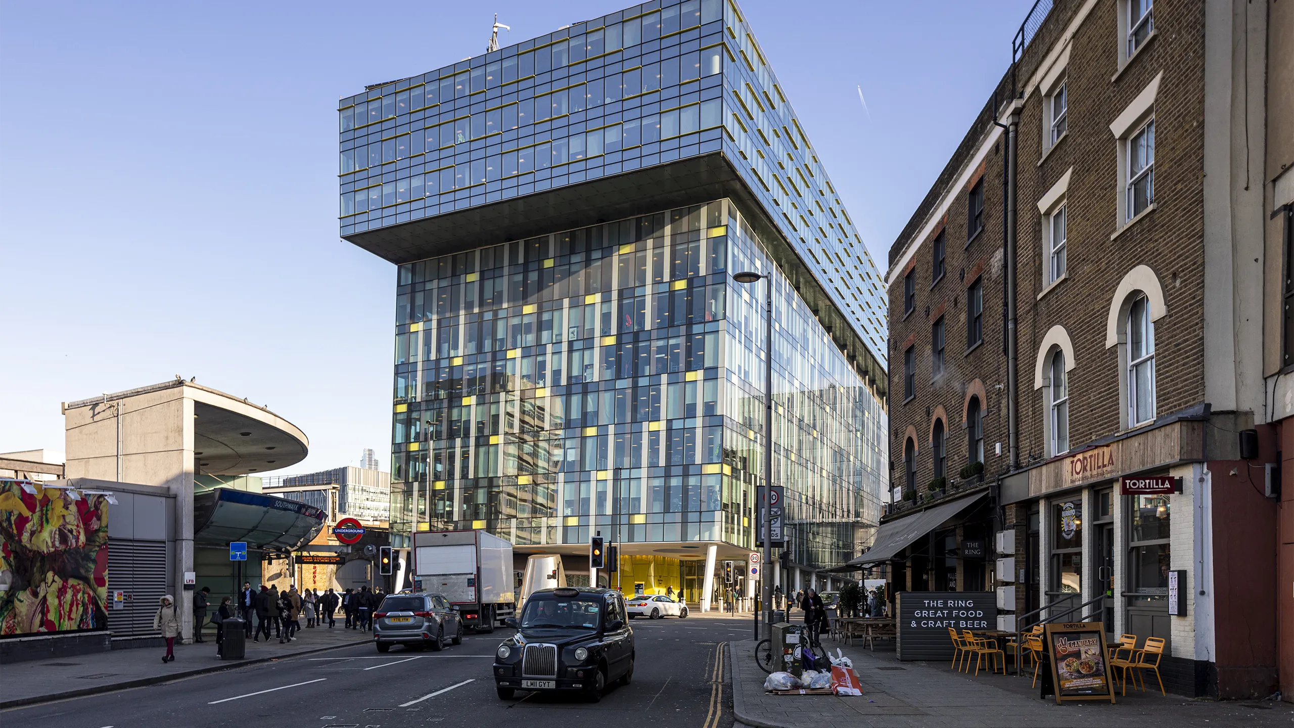 Modern urban street scene in London, featuring a striking contemporary glass building with a cantilevered upper section. This building is set against a backdrop of older brick structures, including a corner restaurant named "Tortilla" with outdoor seating. A classic black London taxi and other vehicles are visible on the street, along with pedestrians walking by. The area appears busy with a mix of architectural styles, reflecting both modern and historical elements of the city. 