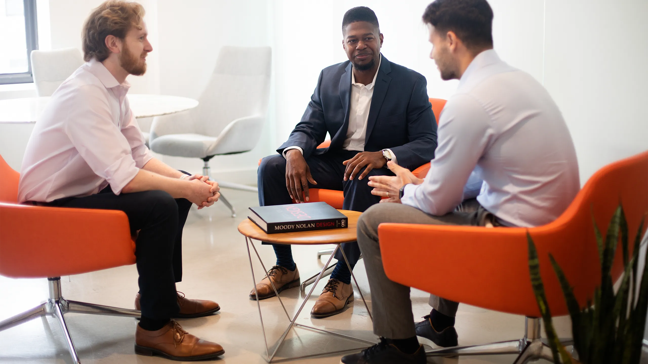 Three male colleagues sitting down on bright orange chairs, engaging in conversation. The office environment is bright and modern with plants behind the chairs.