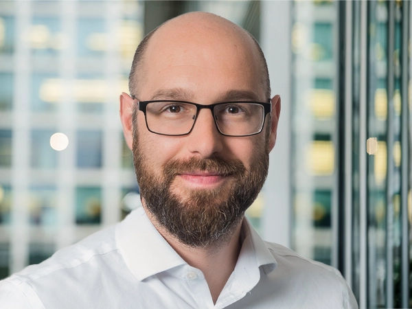 Portrait of Kristoffer Hudson, Associate Director, with city landscape featuring building with lots of windows in the background.