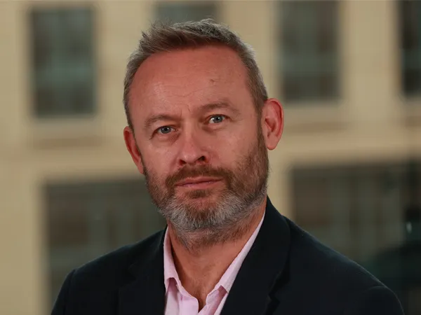 Portrait of Grant Nicolle, Associate Director, UK, standing against brown building in pink shirt and black blazer. 