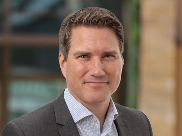 Portrait of Andrew Baker, Europe Lead, outside in front of a blurred brown background featuring a building and mix of natural and urban elements.