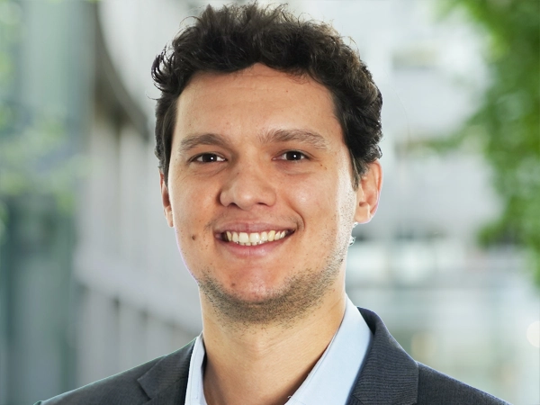 Portrait of Eduardo Carvalho, Senior Project Manager, Real Estate Brazil, standing outside in front of an outdoor urban setting, featuring modern architectural elements with clean lines. There are hints of greenery, suggesting some trees or plants.