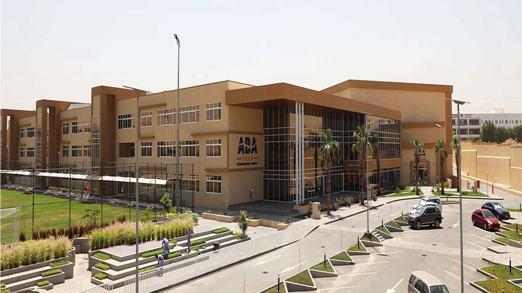 A sand coloured block of school buildings with a car park in front and small playing field to the left of the building. 