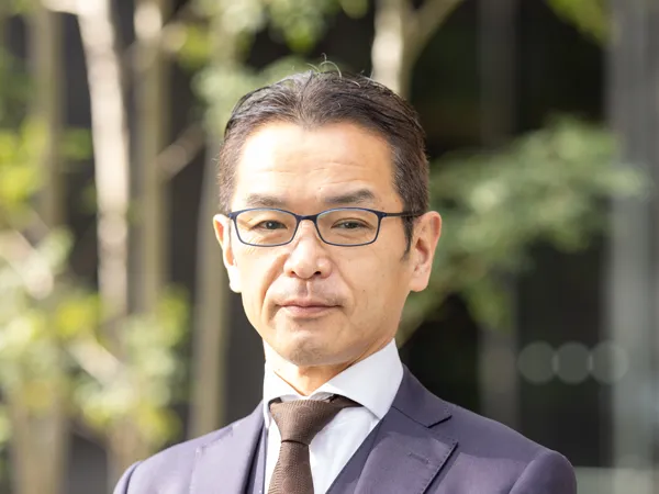Portrait of Yoshihiro Kumano, Senior Project Manager, standing outside in front of tree branches in navy suit.