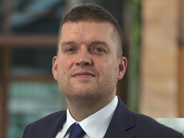 Dan Proud, Director, Infrastructure, UK, outside in front of a blurred brown background featuring a building and mix of natural and urban elements.