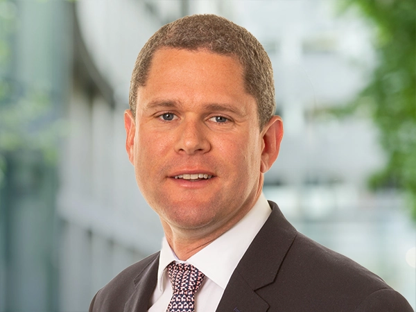 Portrait of Graeme Braithwaite, Director, standing outside in front of an outdoor urban setting, featuring modern architectural elements with clean lines. There are hints of greenery, suggesting some trees or plants.