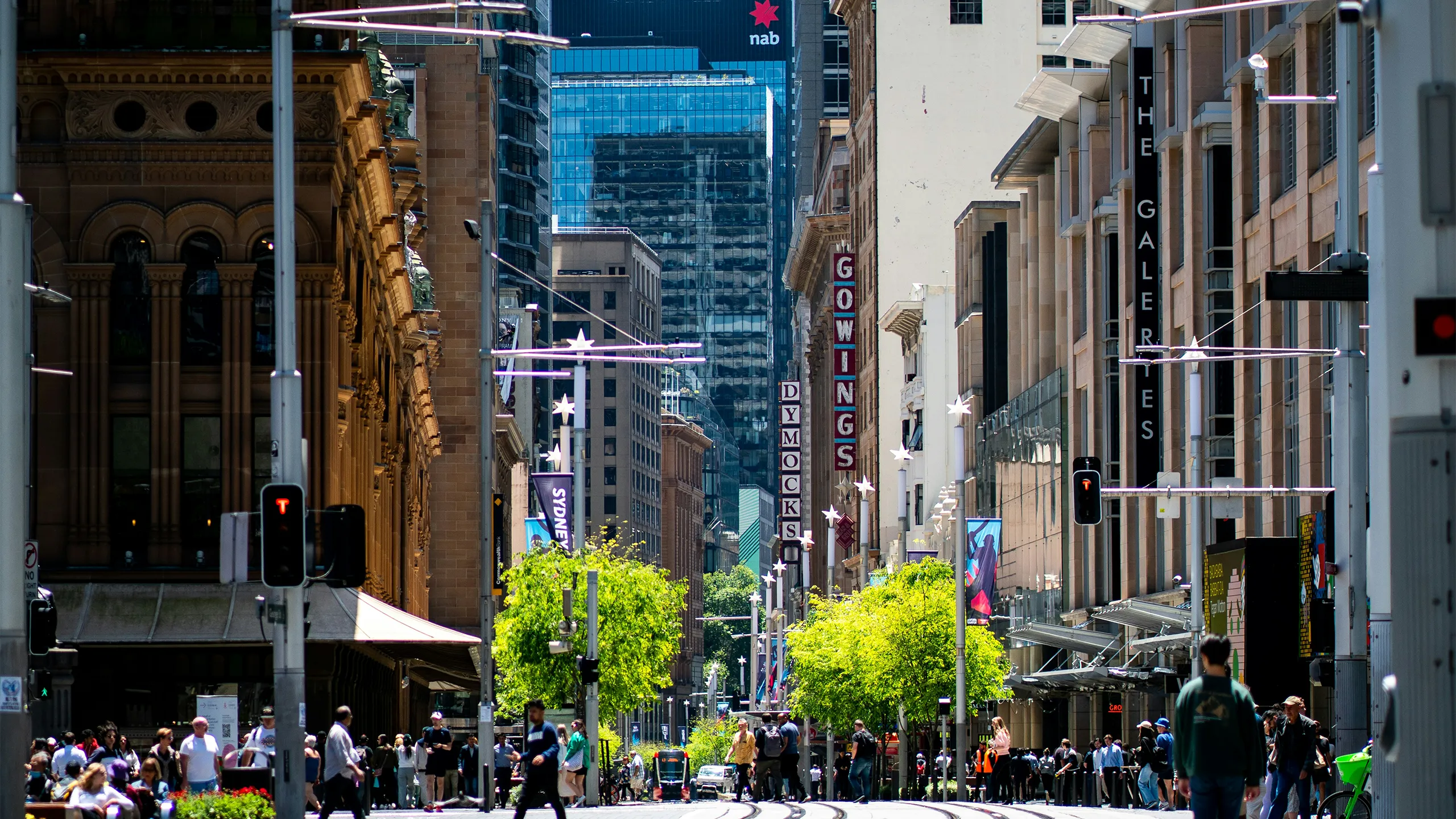 City landscape with people walking and trees in the center with tram coming down in distance.